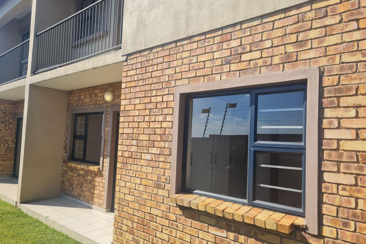 Exterior view of a face-brick apartment building featuring a ground-floor patio, upper-level balcony with metal railings, and dark-framed windows.