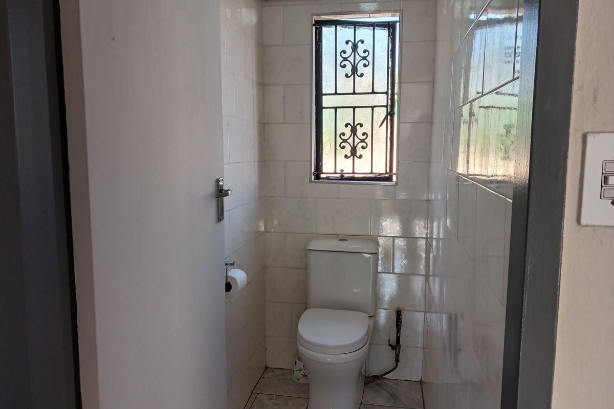 A compact bathroom featuring white wall tiles, a standard toilet, and a window with security bars.