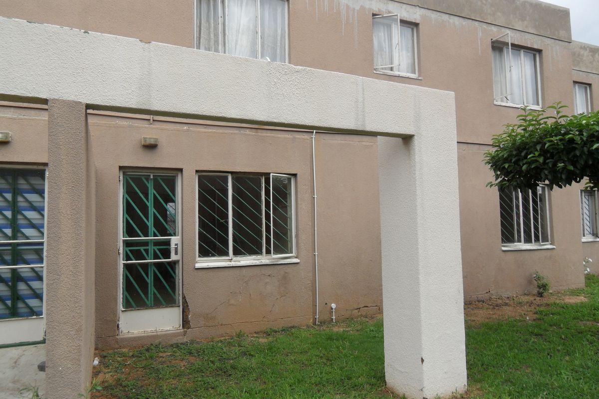 Rear exterior view of a townhouse featuring a concrete pergola structure, metal security gates, and windows with visible signs of weathering and plaster damage.