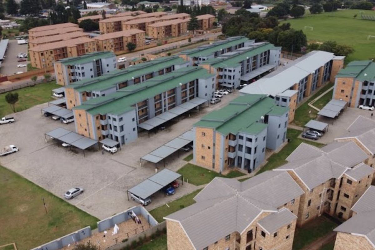 Aerial view of a multi-story apartment complex featuring green metal roofs, paved parking areas with carports, and landscaped grounds.