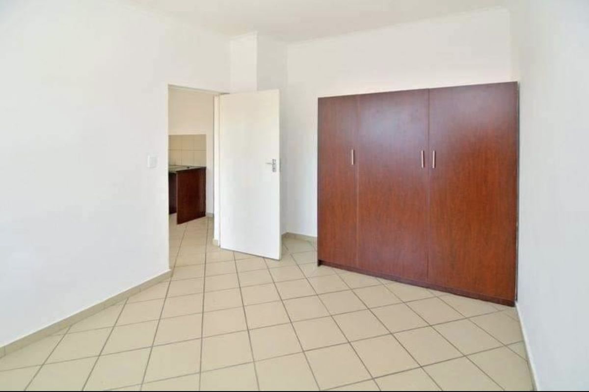 A bedroom featuring neutral tiled flooring and a built-in wooden wardrobe, with a doorway leading to an adjacent kitchen area.