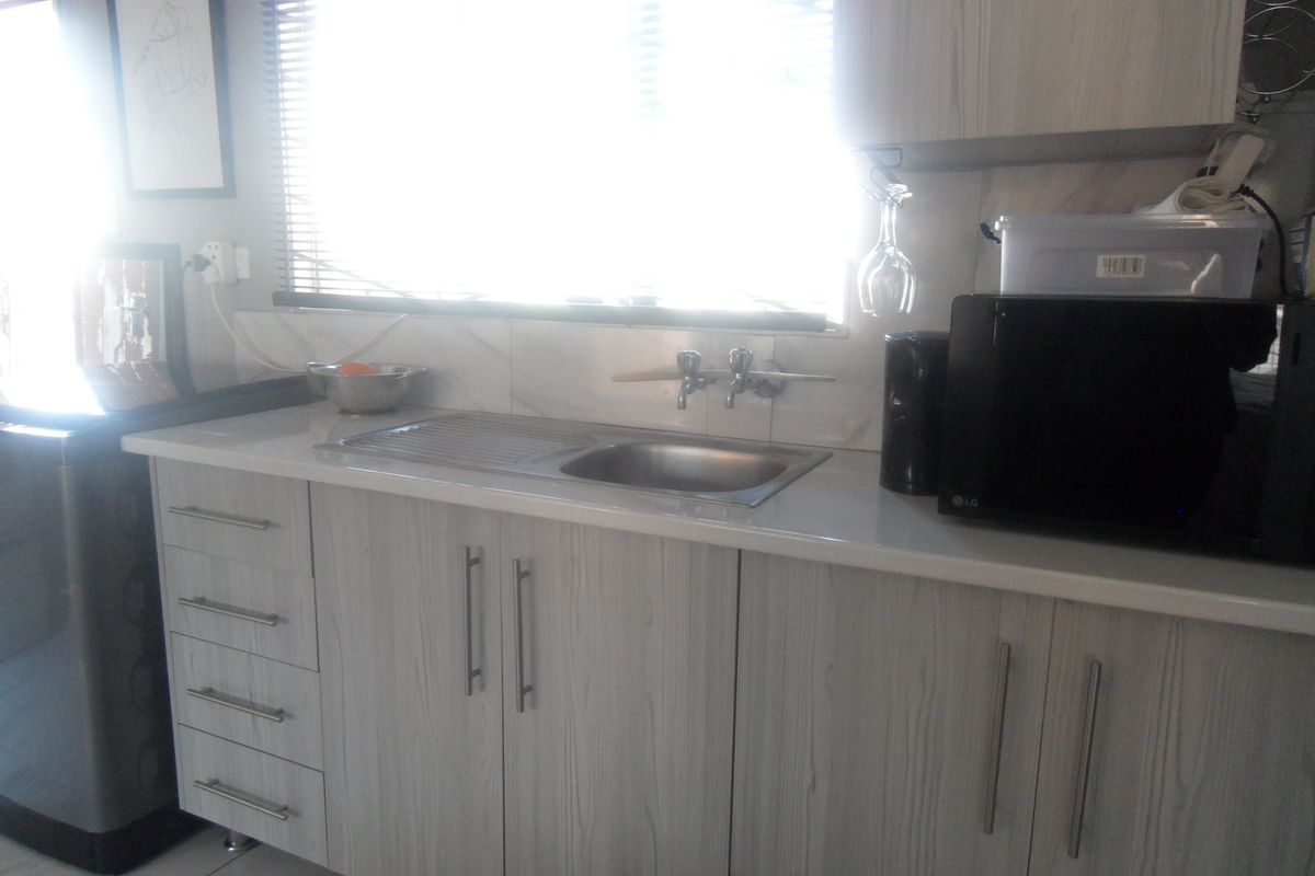 A functional kitchen area featuring light wood-grain cabinetry, a stainless steel sink, and a tiled backsplash under a window with blinds.