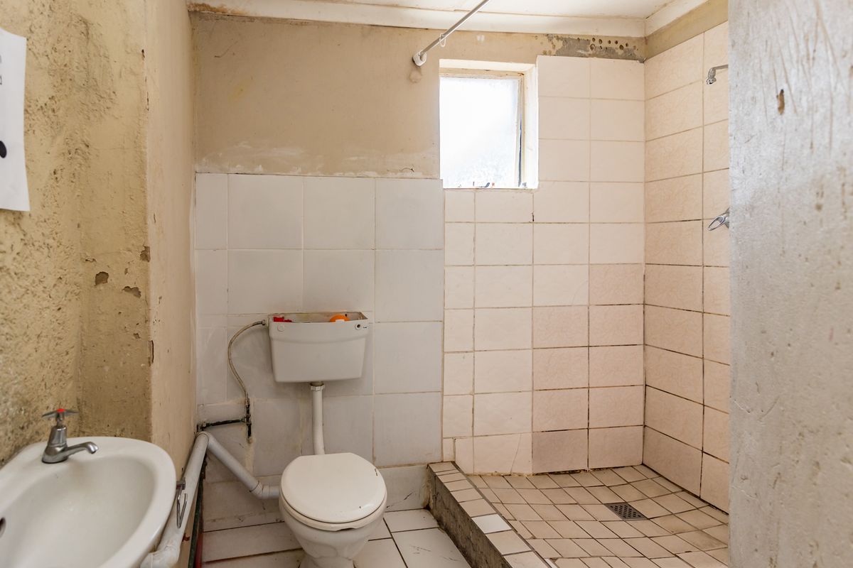 A bathroom in need of significant renovation, featuring a basic toilet, pedestal basin, and a tiled shower area with visible wall damage.