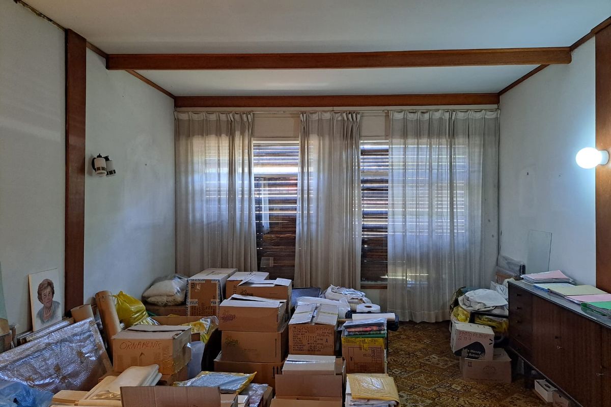 A living room space featuring timber ceiling beams, large windows with curtains, and carpeted flooring, currently used for storage.