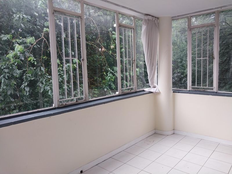 An enclosed balcony or sunroom featuring tiled flooring, large windows with security bars, and a structural column, offering a bright space with garden views.