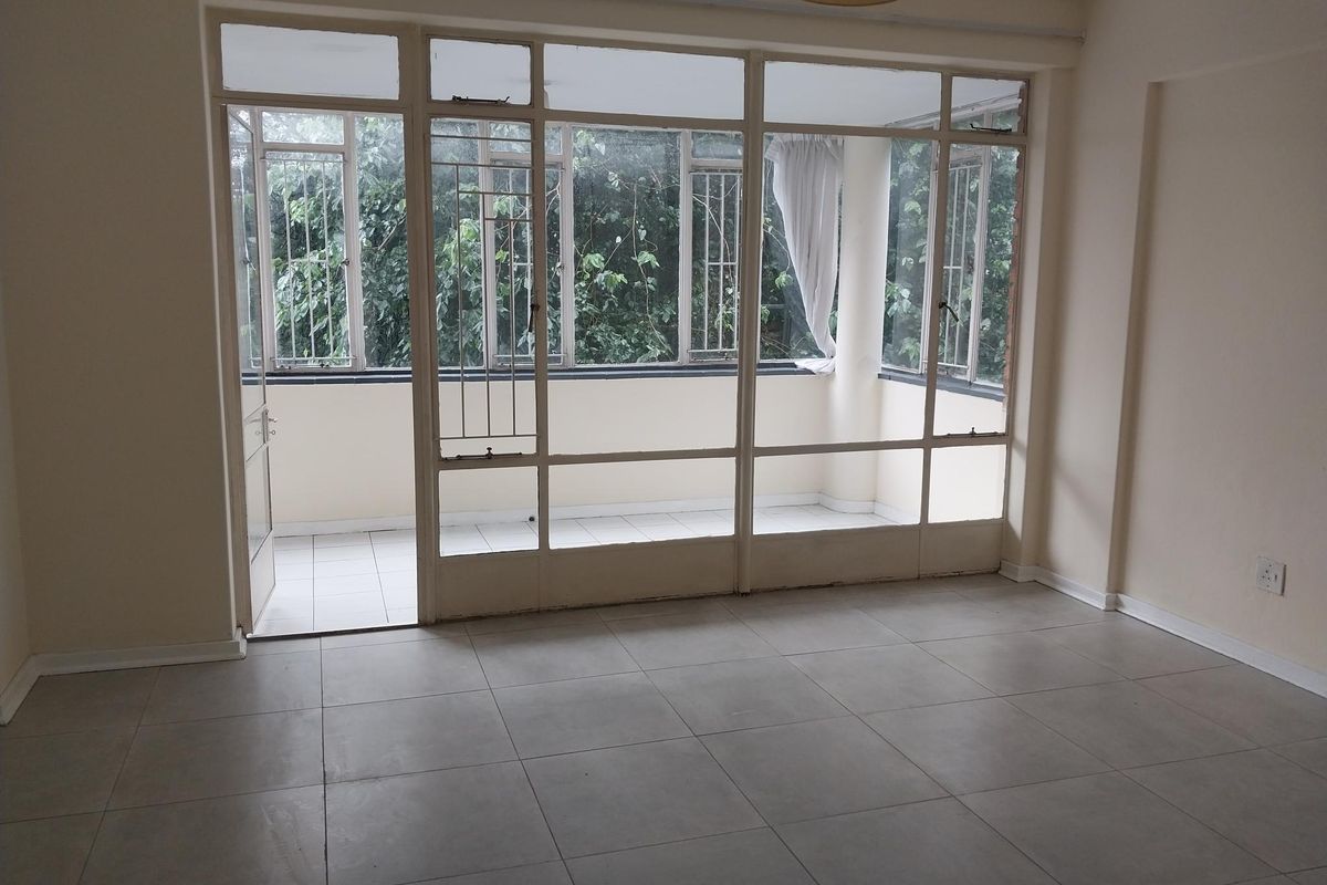 An empty living room featuring large windows leading to an enclosed balcony, with neutral tiled flooring and ample natural light.