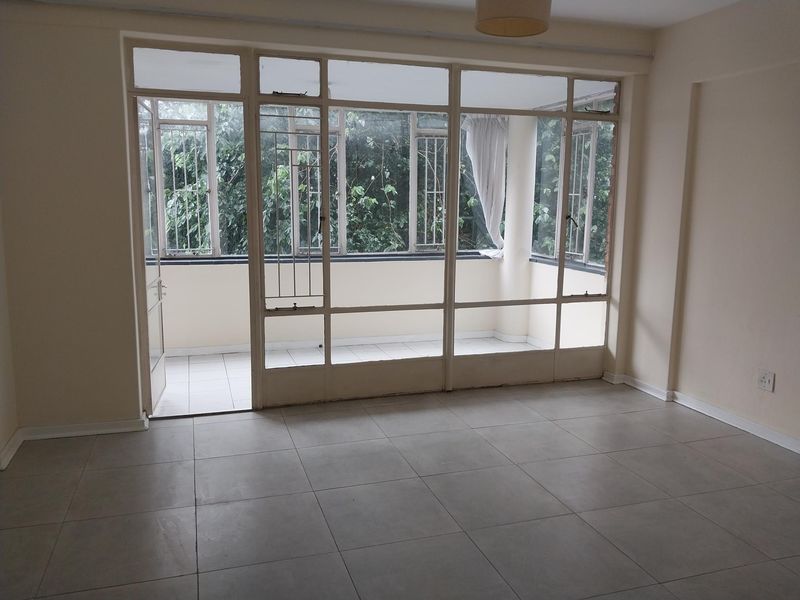 An empty living room featuring large windows leading to an enclosed balcony, with neutral tiled flooring and ample natural light.