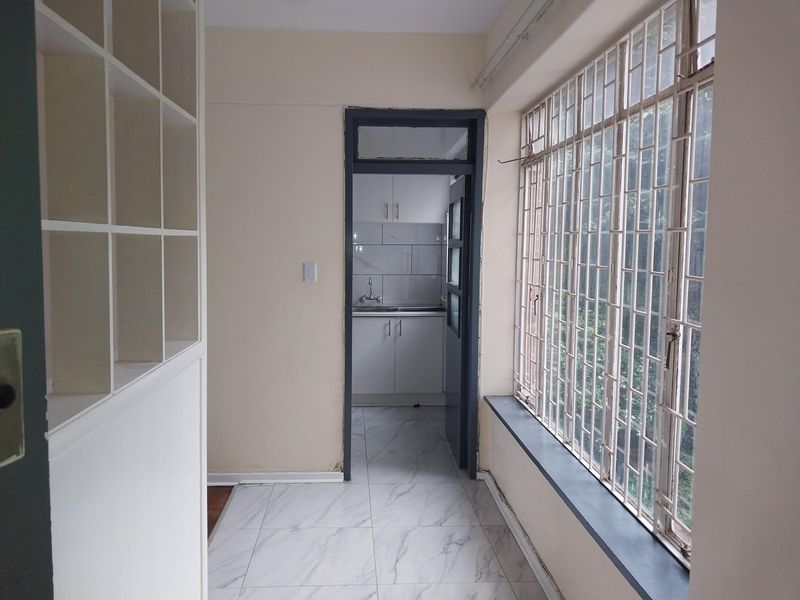 A bright hallway featuring tiled flooring, built-in shelving, and security bars on the window, leading through to a kitchen area.
