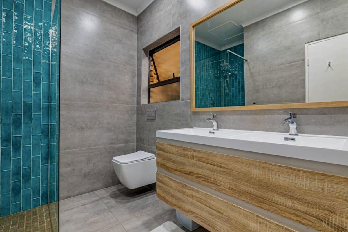 Modern bathroom featuring a double vanity with wood-look cabinetry, wall-mounted toilet, and striking teal subway tile accents against grey porcelain wall tiles.