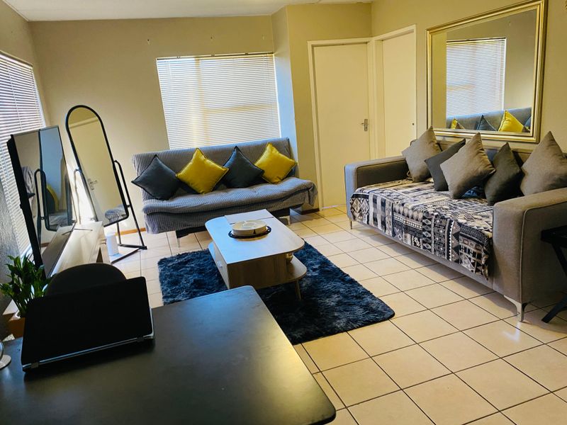 A functional living room featuring tiled flooring, neutral wall tones, and large windows fitted with venetian blinds.