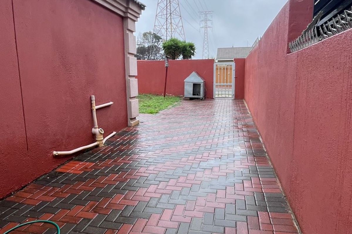 A paved side walkway featuring high perimeter walls, a metal security gate, and exposed exterior plumbing fixtures.