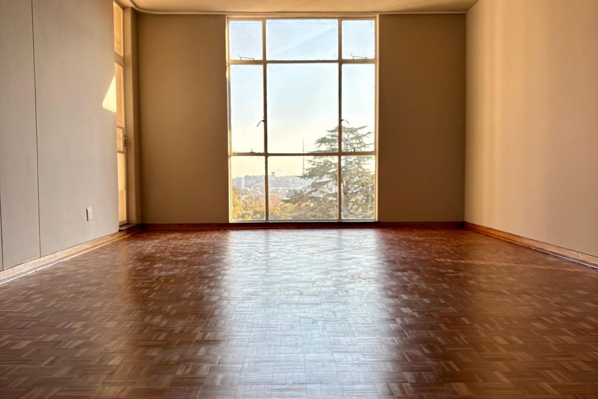 A spacious, empty living area featuring classic parquet flooring, large windows with natural light, and elevated views.
