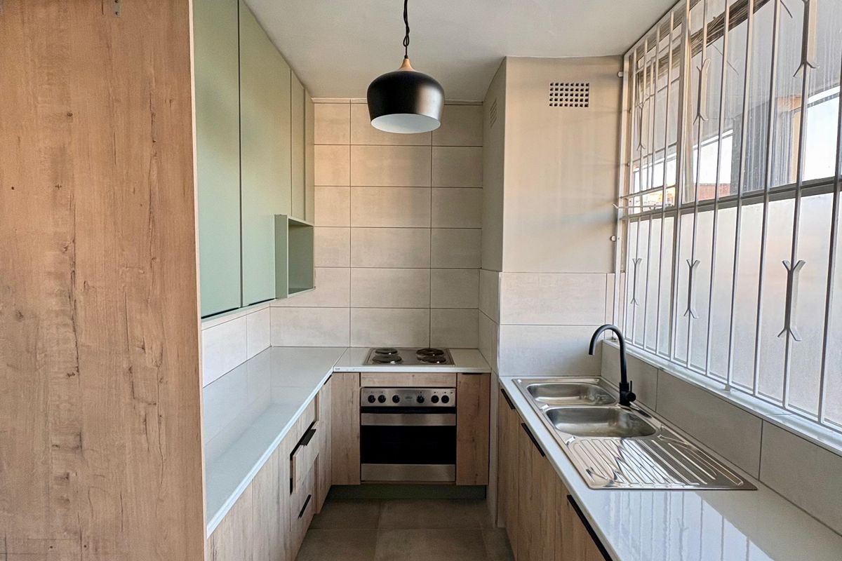A modern galley-style kitchen featuring light wood-grain cabinetry, stone-look countertops, a built-in oven and hob, and a double stainless steel sink.