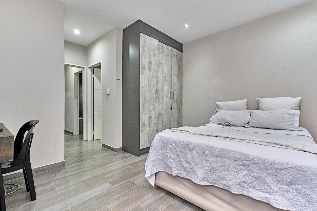 A modern bedroom featuring built-in cabinetry with a wood-grain finish, recessed ceiling lighting, and neutral tiled flooring, leading to an adjacent hallway.