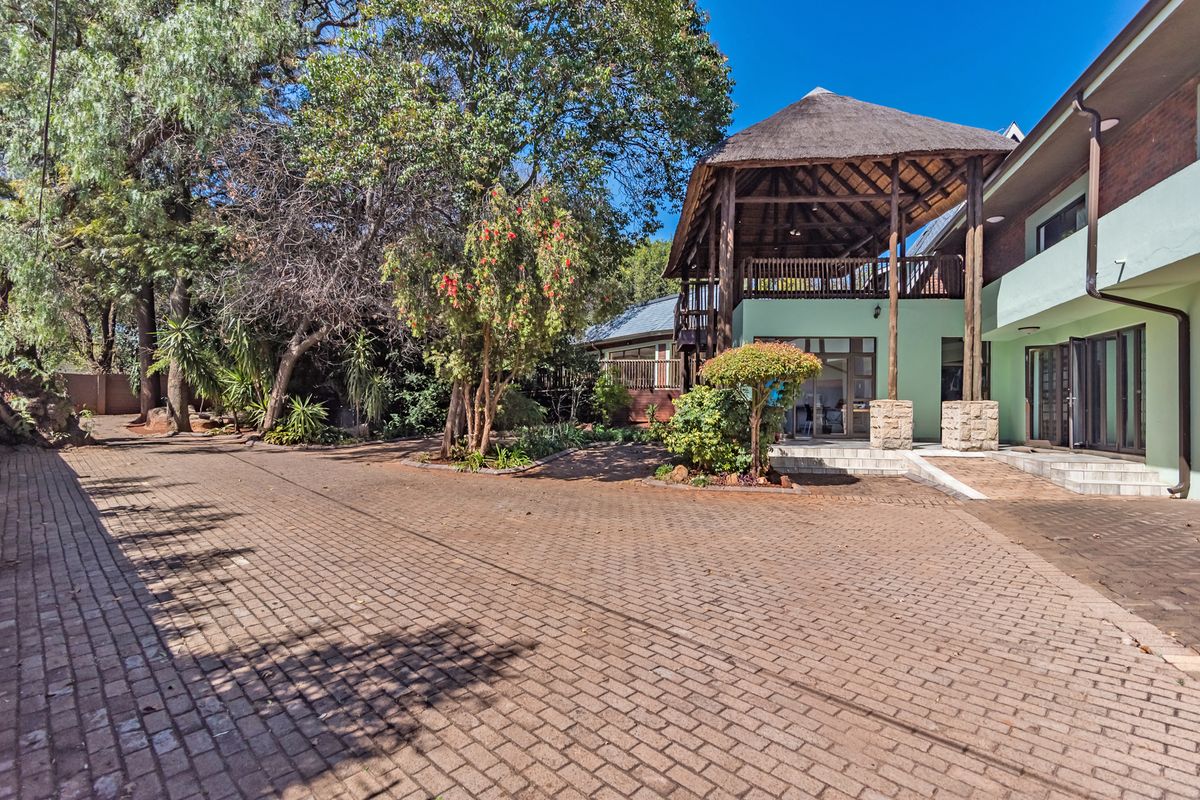 A spacious paved driveway leading to a multi-level home featuring a thatched roof patio and established garden surroundings.