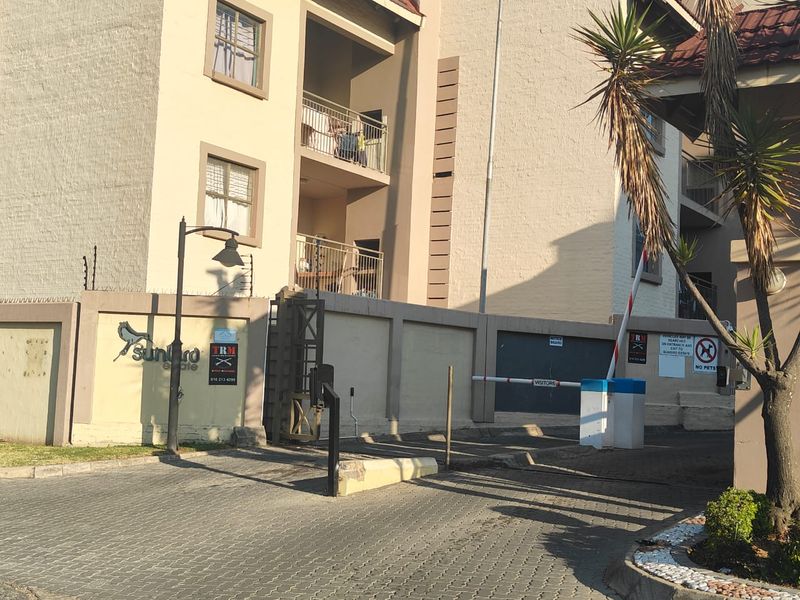 Front entrance view of the apartment complex featuring a paved driveway, security boom gate, perimeter wall with electric fencing, and multi-story residential building facade.