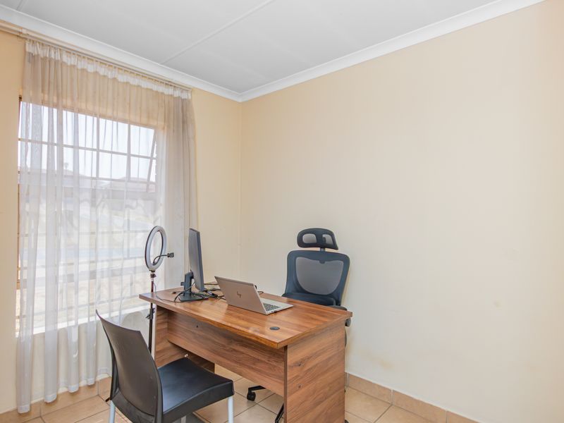 A functional home office space featuring tiled flooring, neutral wall finishes, and natural light provided by a large window with sheer curtains.