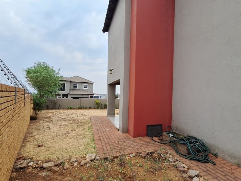 Rear exterior view showing a paved walkway, covered patio area, brick boundary wall with electric fencing, and a large side garden space.