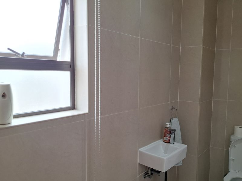 A compact, modern bathroom featuring neutral wall tiles, a wall-mounted basin, and a toilet, with a functional casement window for ventilation.
