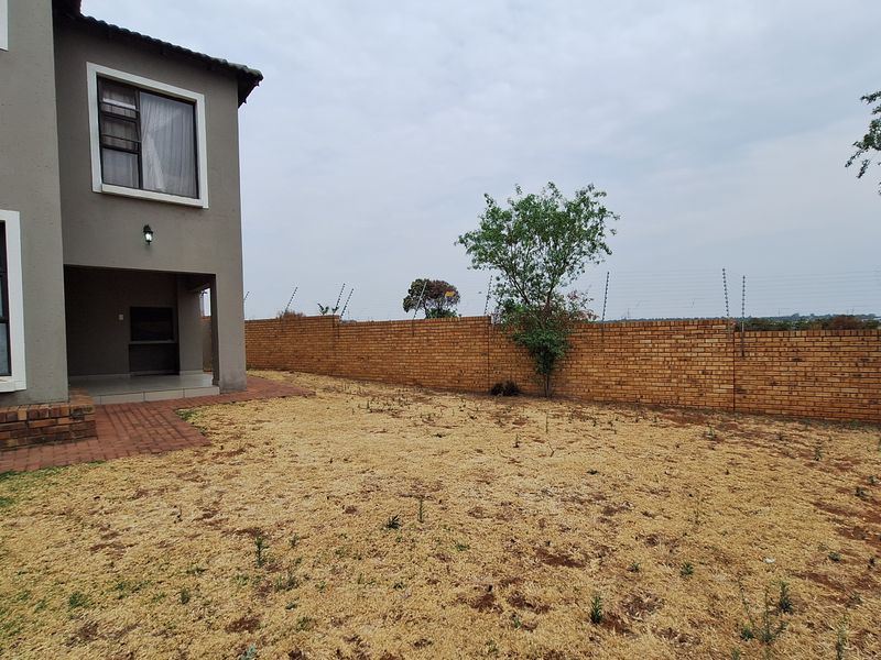 Rear view of the property featuring a covered patio with a built-in braai, paved walkway, and a large garden enclosed by a brick boundary wall.