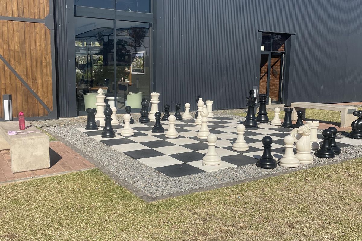 Outdoor area featuring a large-scale chess board set against a modern industrial-style building with corrugated metal cladding and timber accents.