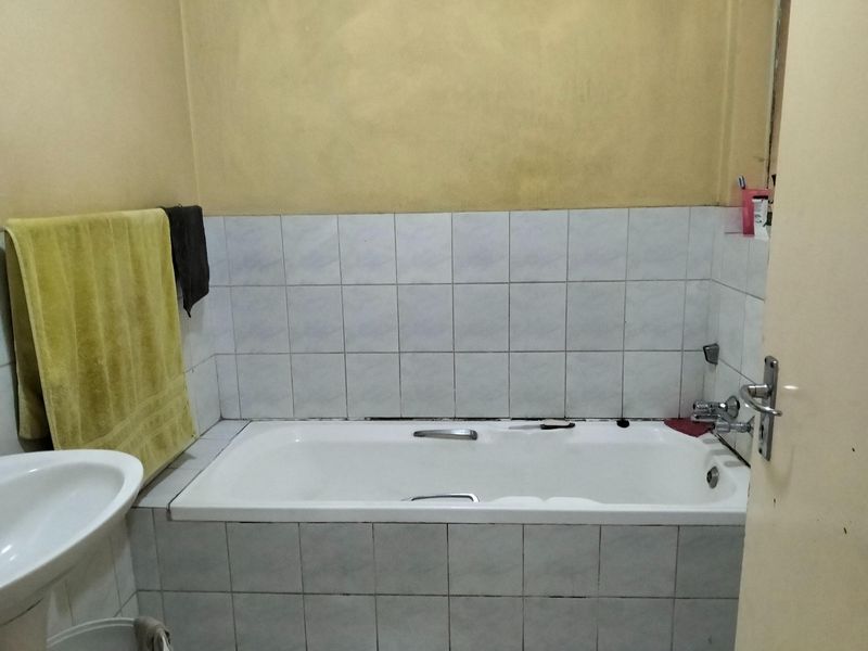 A functional bathroom featuring a built-in bathtub and wall-mounted basin with tiled splashbacks, requiring cosmetic updates to the wall finishes and grout.