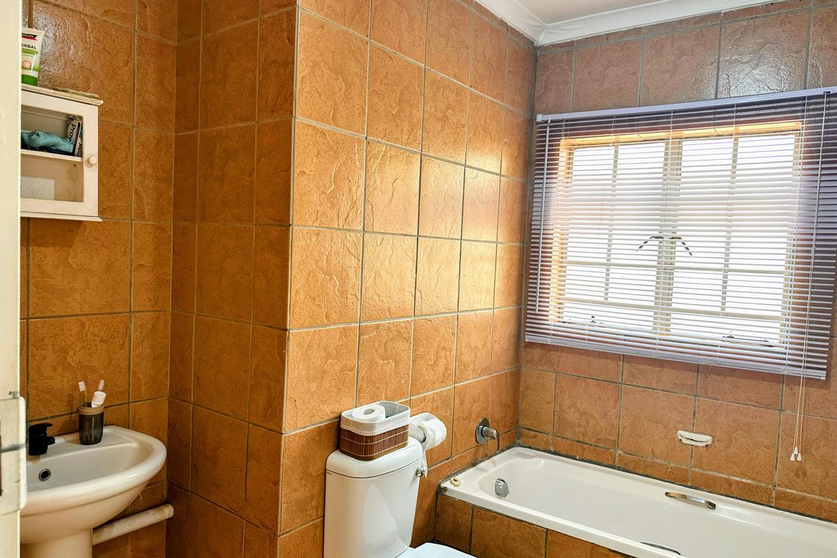 A dated bathroom featuring full-height terracotta-style wall tiles, a standard bathtub, a pedestal basin, and a window with fitted blinds.