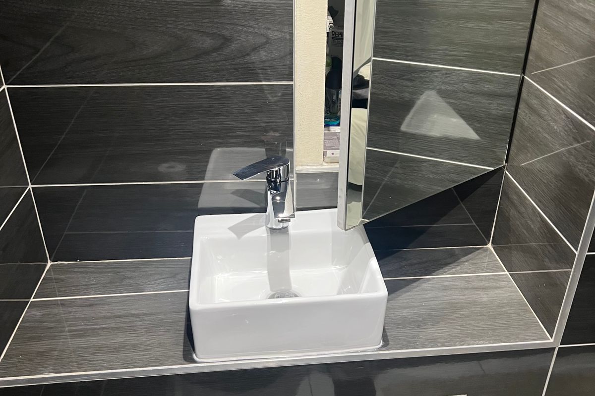 A modern bathroom vanity featuring a square vessel sink, chrome faucet, and dark wood-look tiled splashback with integrated storage.