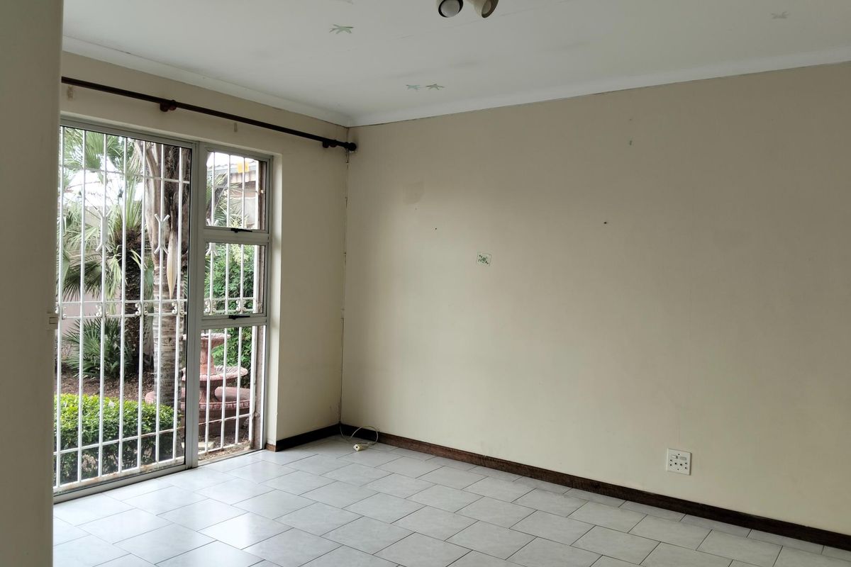 An empty living room featuring tiled flooring, neutral walls, and a sliding glass door with security bars providing access to the garden.