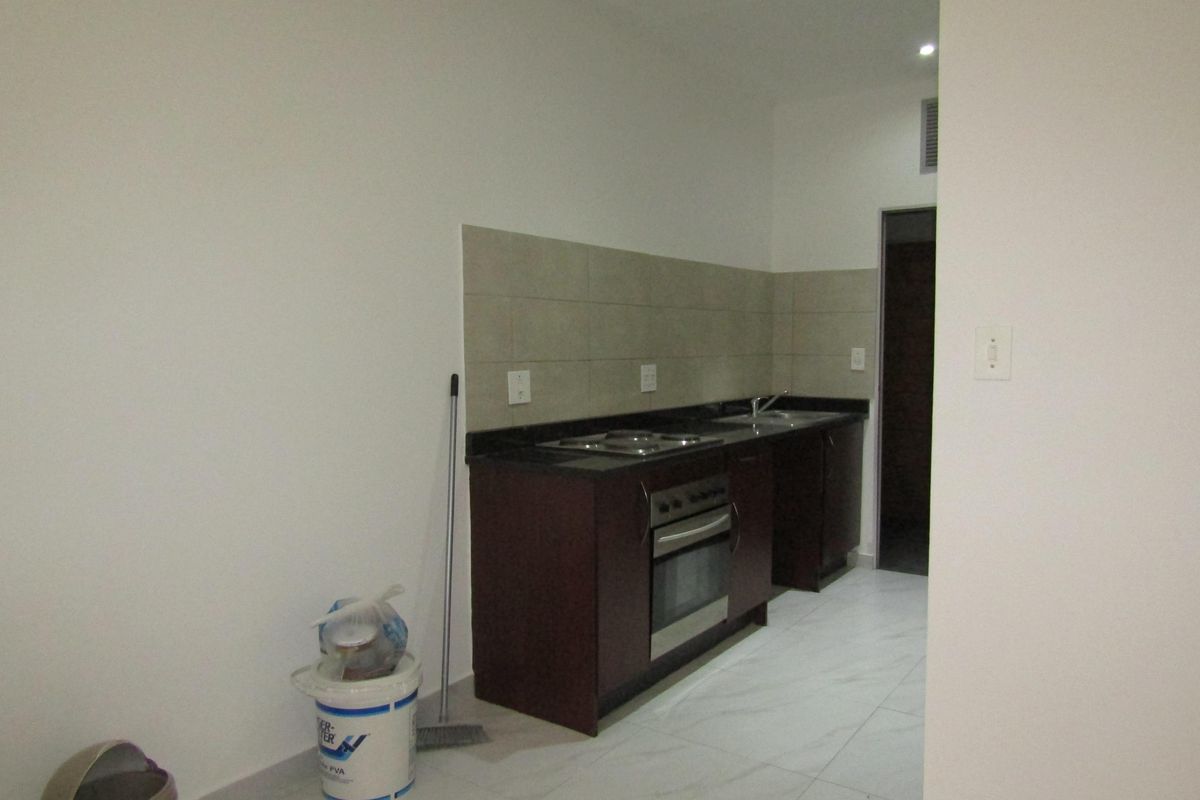 A compact kitchen featuring dark cabinetry, a built-in oven and hob, stone countertops, and tiled splashbacks, set within a clean, tiled open-plan space.