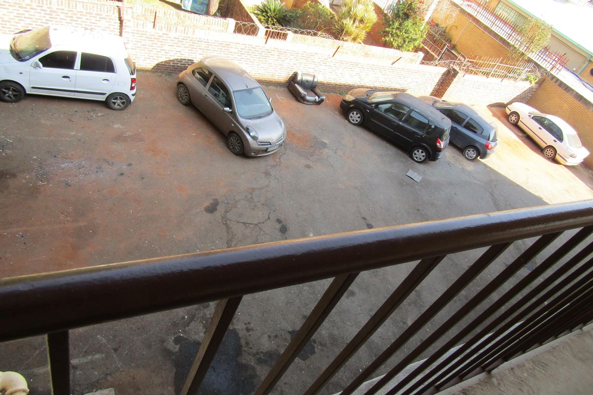 An elevated view of a shared asphalt parking area enclosed by a brick boundary wall, viewed from a balcony with a metal railing.