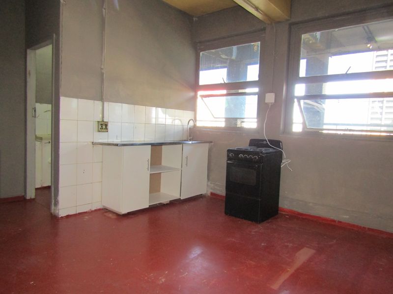A basic, unfinished kitchen area featuring a small sink unit, tiled splashback, and a freestanding stove, requiring significant renovation and cosmetic updates.
