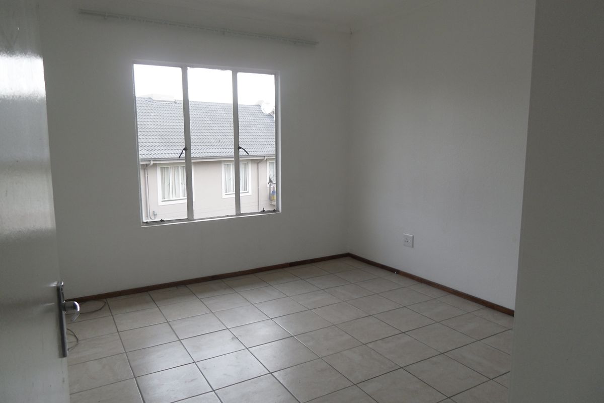 A bright, empty bedroom featuring neutral ceramic floor tiles, white walls, and a large window providing natural light and a view of the complex.