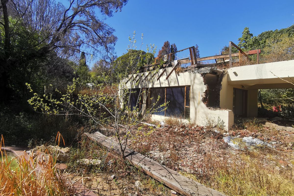Rear view of a derelict residential property featuring a damaged structure, collapsed roof elements, and an overgrown garden area with a swimming pool.