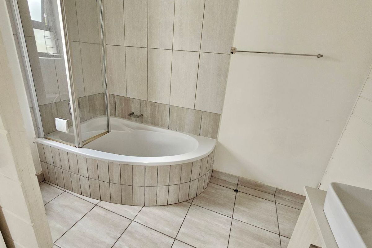 A functional bathroom featuring a built-in bathtub with a glass shower screen, tiled surround, and neutral floor tiling.