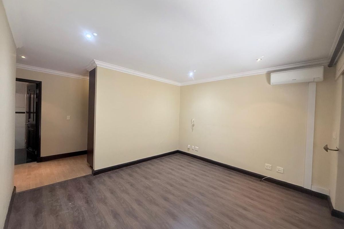 A spacious, empty living area featuring modern wood-look flooring, recessed ceiling lighting, crown molding, and a wall-mounted air conditioning unit.