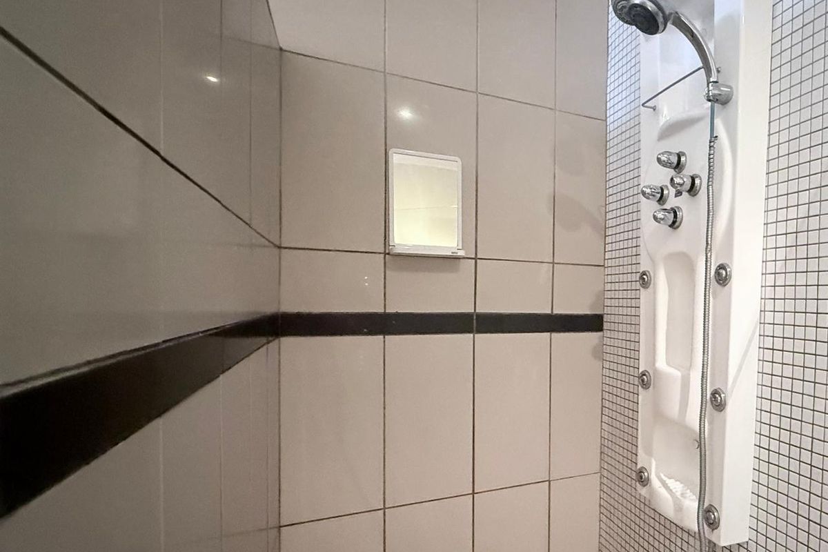 A shower enclosure featuring a modern multi-jet shower panel, tiled walls with a decorative border, and a small built-in wall mirror.