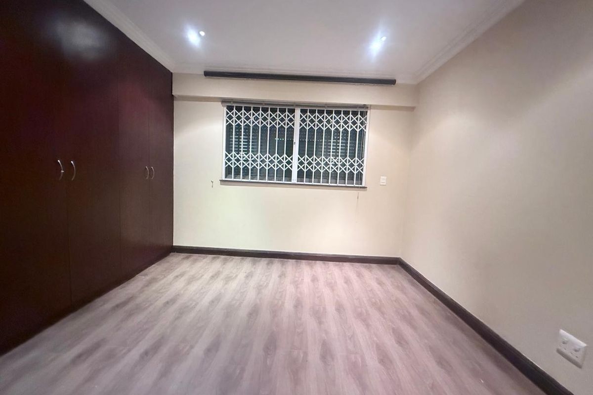 A clean, empty bedroom featuring wood-look laminate flooring, built-in dark wood cabinetry, and a window secured with a white metal security gate.