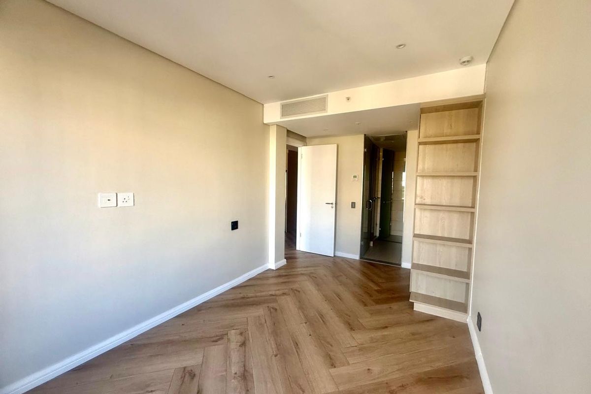 A bright, empty bedroom featuring modern herringbone wood flooring, built-in shelving, and integrated ceiling air conditioning.