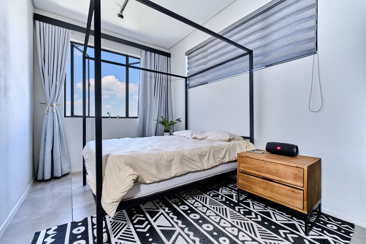 A bright bedroom featuring large windows with modern blinds, neutral tiled flooring, and clean white walls, offering a contemporary and airy living space.