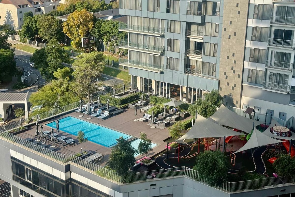 Elevated aerial view of a modern apartment complex featuring a rooftop swimming pool, sun deck, and a landscaped children's play area.