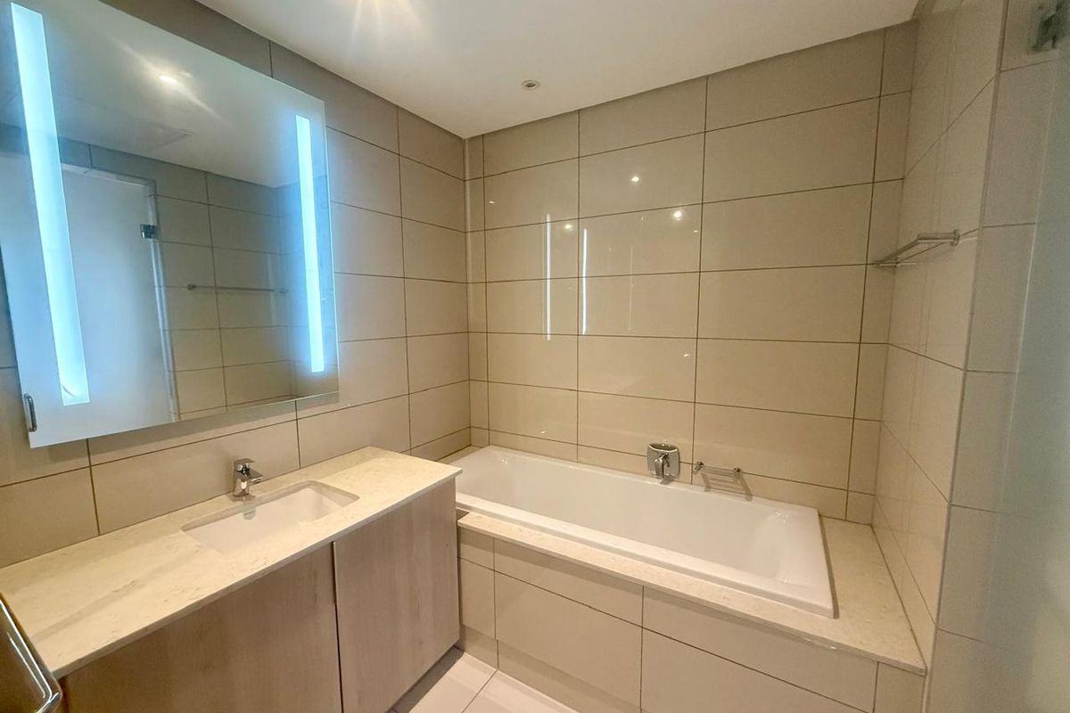 Modern bathroom featuring a built-in bathtub, vanity with integrated sink, and a large illuminated mirror with tiled walls and flooring.