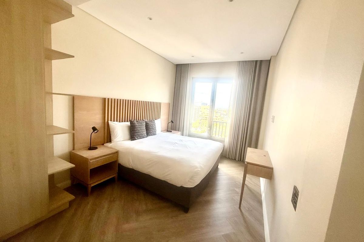 A modern bedroom featuring light wood herringbone flooring, a built-in headboard with integrated shelving, and floor-to-ceiling curtains.