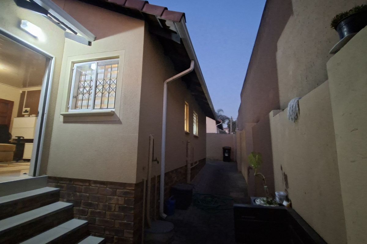 A side exterior view of the house showing a paved walkway, boundary wall, and security features on the windows.