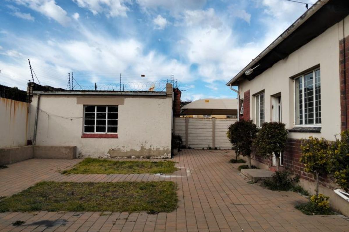 Rear exterior view of the property featuring a paved courtyard, small garden patches, and a detached outbuilding with security fencing.