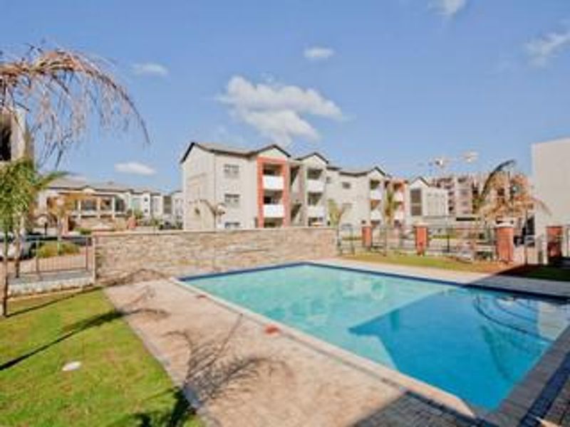 A well-maintained communal swimming pool area featuring a stone-clad feature wall, paved decking, and surrounding landscaped garden within an apartment complex.