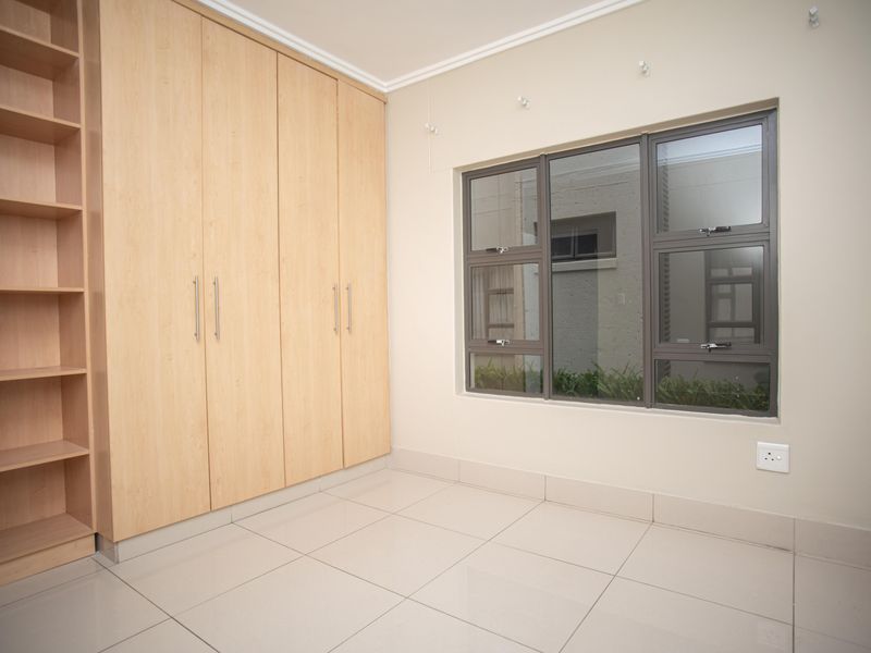 A clean, empty bedroom featuring light wood-finish built-in cupboards, neutral tiled flooring, and modern aluminium-framed windows.