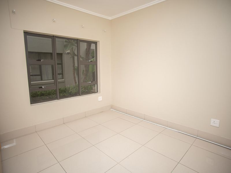 An empty bedroom featuring neutral tiled flooring, a built-in cupboard, and an aluminium-framed window overlooking an exterior courtyard.