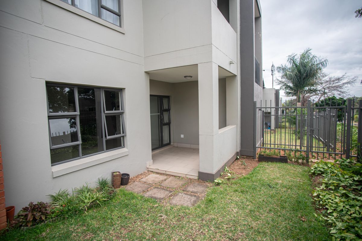 A ground floor apartment exterior featuring a covered tiled patio, aluminium window frames, and a small private garden area with metal perimeter fencing.