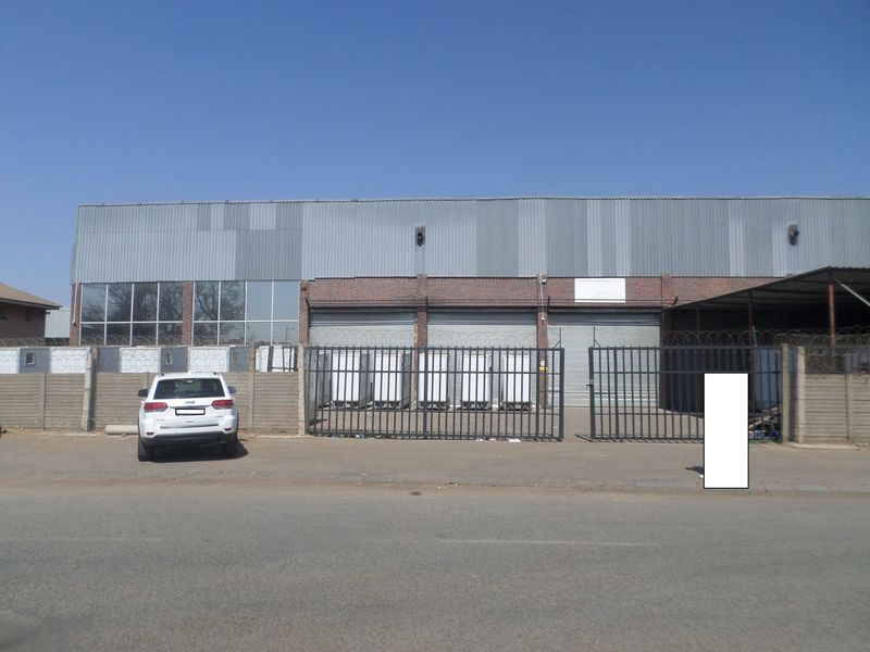 Front elevation of an industrial warehouse facility featuring corrugated metal cladding, brickwork, multiple roller shutter doors, and secure perimeter fencing.